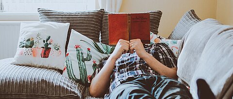 Eine Person liegt lesend auf der Couch. Die Füße zeigen in Richtung Kamera, das Gesicht ist vom Buch verdeckt.