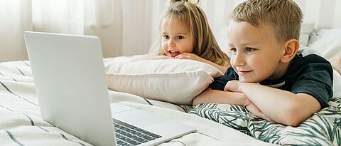 Zwei Kinder liegen auf dem Bett und schauen auf einen Laptopbildschirm